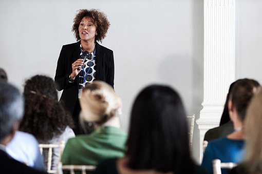 Dans une salle de conférence, une femme debout tient un micro et parle à une assistance composée d'adultes assis, vus de dos.