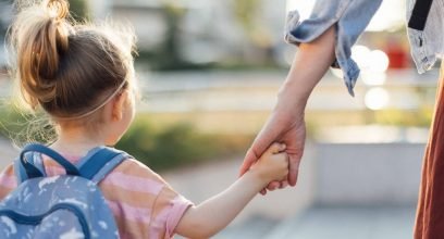 Une petite fille de maternelle, sac sur le dos, vue de dos, tient la main d'une femme adulte dont on voit le bras et un bout de jupe