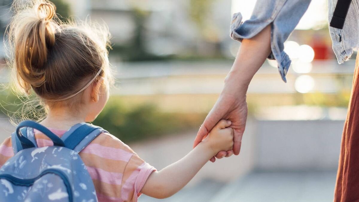 Une petite fille de maternelle, sac sur le dos, vue de dos, tient la main d'une femme adulte dont on voit le bras et un bout de jupe
