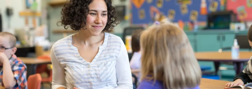 salle de classe de primaire, une enseignante sourit face à une élève vue de dos